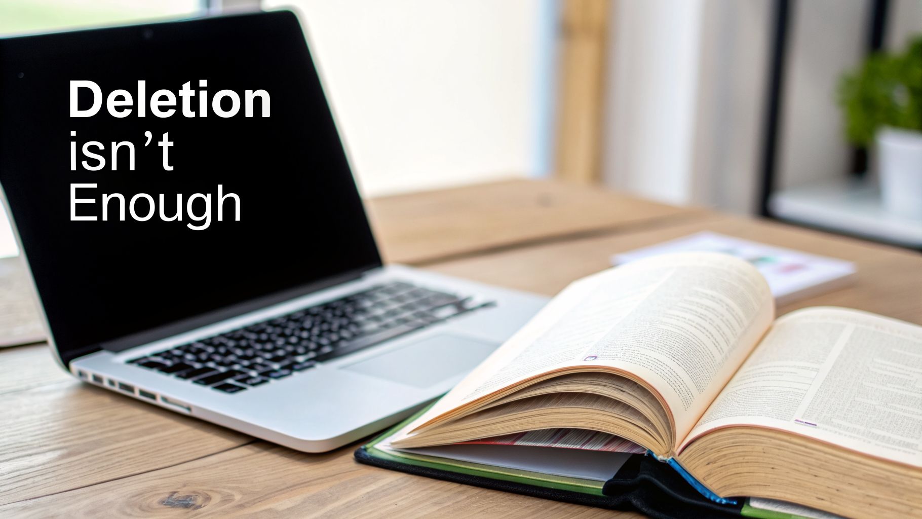 A laptop displaying 'Deletion isn't Enough' on screen, with an open book on a wooden desk.