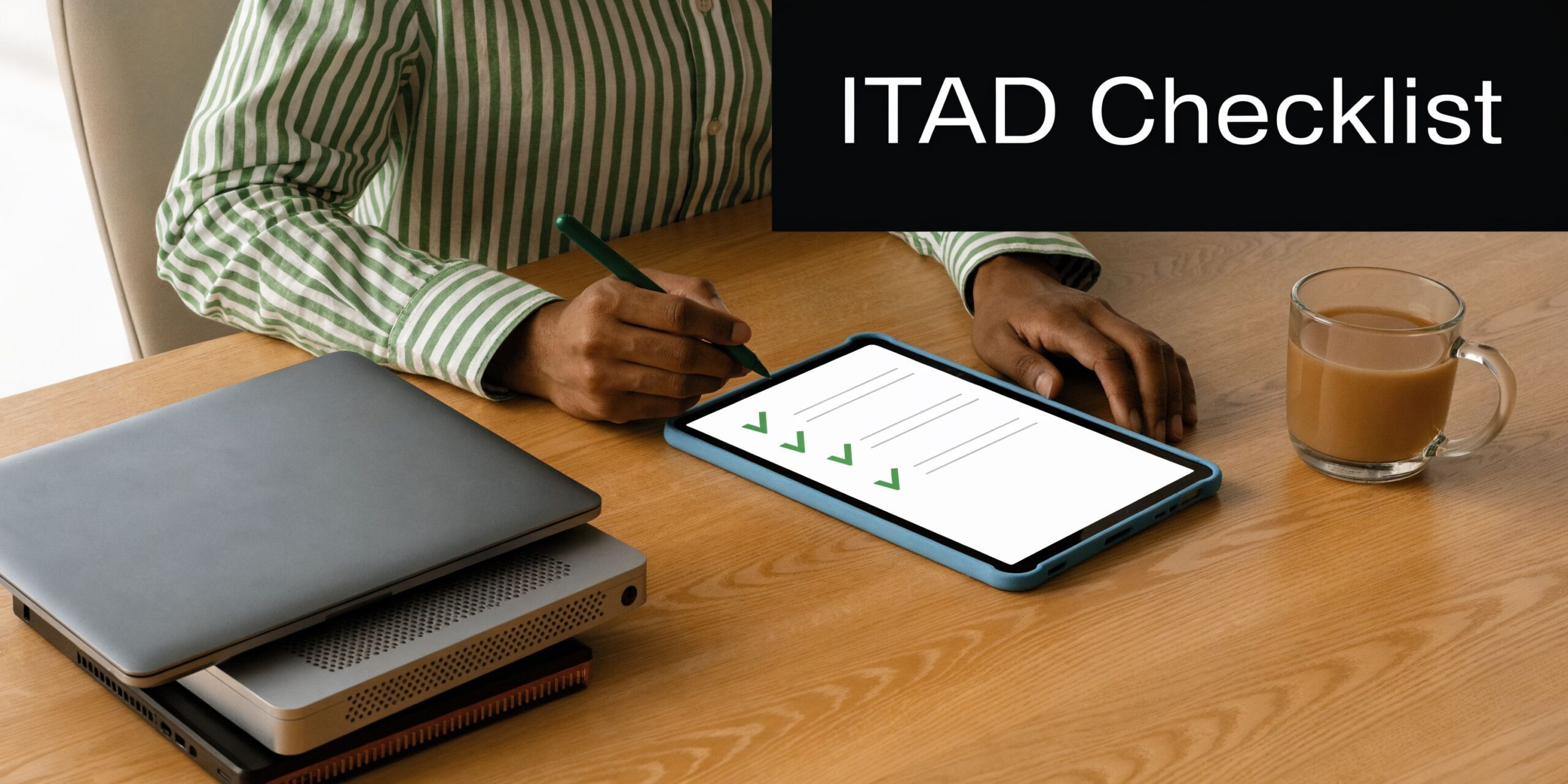 A person writing on a tablet with an ITAD checklist displayed at a desk with stacked laptops.